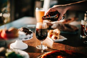 Sunlit scene with hand pouring dark beer into a glass. Includes baked goods and desserts on table.